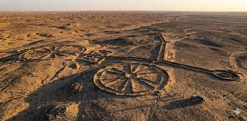Fotografia aérea de grandes estruturas de pedra pré-históricas em forma de roda em um deserto rochoso, com um grupo de pesquisadores e um veículo 4x4 no canto inferior para escala.